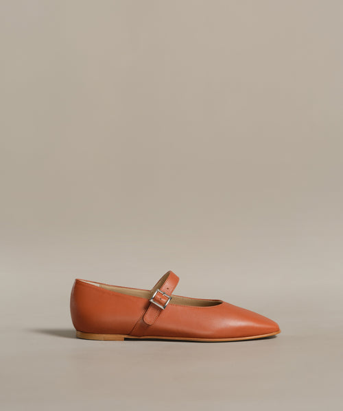 The Lana Flat is an orange-brown leather Mary Jane shoe featuring a tapered toe and a thin strap with a small buckle across the instep, shown on a neutral background.