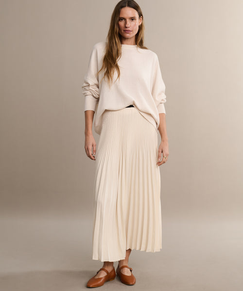 A woman with long, wavy hair poses casually against a beige background, wearing a loose cream sweater, a pleated cream skirt, and the Lana Flat—a brown Mary Jane shoe with a tapered toe that adds feminine flair.