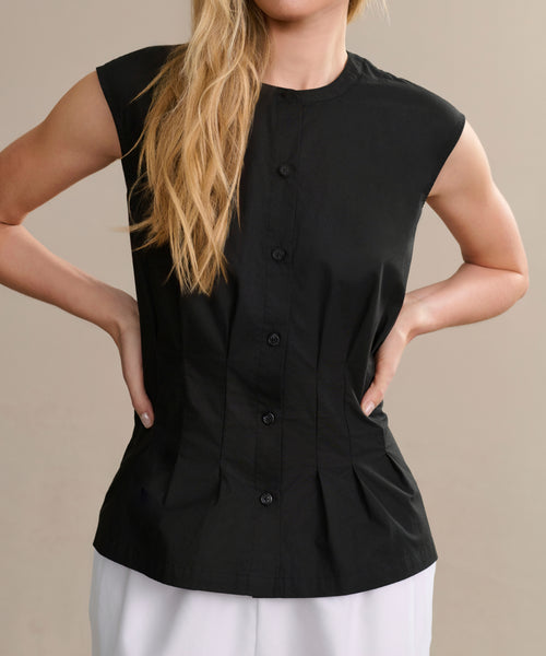 A woman with long blonde hair poses with hands on hips, wearing the Lara Top—a sleeveless black button-front blouse—paired with white pants against a beige background.