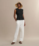A woman wears the Lara Top—a sleeveless black button-front style—paired with white loose pants, black sandals, and a wide-brimmed straw hat, standing against a plain beige background.