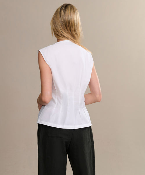 A person with long blonde hair, seen from behind, wears the sleeveless white Lara Top and loose black pants against a plain beige background.
