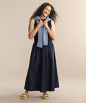 A woman with long curly hair smiles in a sleeveless navy top and matching Lauren Skirt, a light blue sweater draped over her shoulders, tan sandals on her feet. She stands against a plain beige background.