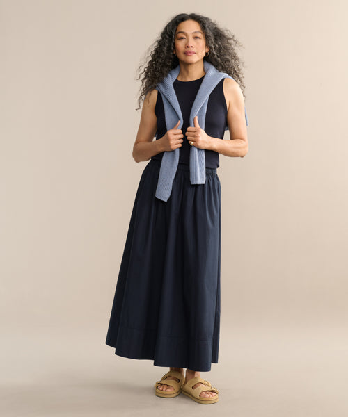 A woman with long curly hair stands against a beige background, wearing a sleeveless navy top, the Lauren Skirt in matching navy, tan sandals, and a light blue sweater draped over her shoulders.