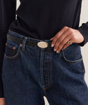 A person wears a black long-sleeve shirt tucked into high-waisted blue jeans, accessorized with the Leather Audrey Belt featuring a brass buckle, resting one hand on their hip and wearing a gold ring.