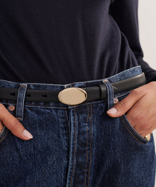 A person in a black long-sleeve top and blue jeans wears the Leather Audrey Belt—calf leather with a gold oval buckle—hands casually tucked into their front pockets.