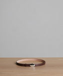 The Leather Audrey Belt, crafted in Italy from brown calf leather, is displayed in a circular shape on a light wooden surface with a gray background.
