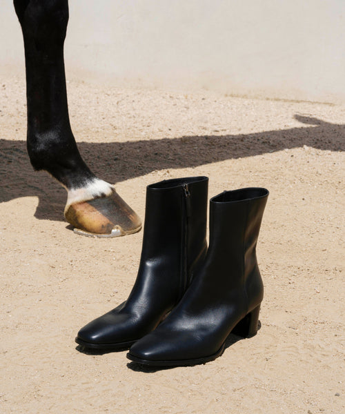 The Leather Bobby Boot, crafted from black Italian leather with block heels, is pictured on sandy ground beside a horse’s front leg and hoof against a plain, light-colored background.