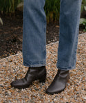 A person in blue jeans wears the versatile Leather Bobby Boot for women, standing on a gravel path with green plants nearby.