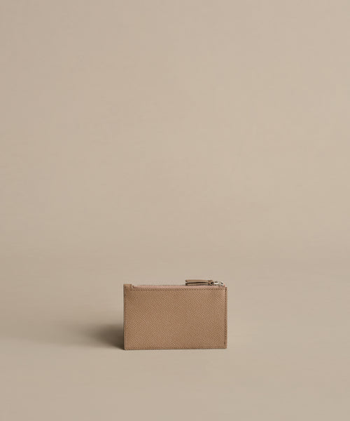 The Leather Card Holder, in taupe with a zippered closure, is centered on a beige background for a minimalist, monochromatic look—ideal for anyone who prefers to travel light.