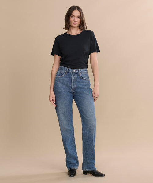 A person with shoulder-length brown hair wears a black t-shirt, high-waisted blue jeans, and the Leather Carmine Boot, standing against a plain beige background.