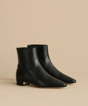 The Leather Carmine Boot, a pair of black leather ankle boots with low heels, pointed toes, and side zippers, showcases a sleek minimalist design against a beige background.