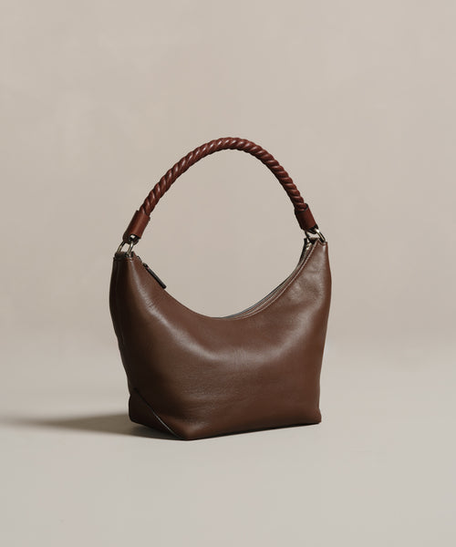 The Crescent Shoulder Bag, handmade in Italy from soft brown leather, showcases a curved silhouette and a thick braided handle, set against a plain beige background.