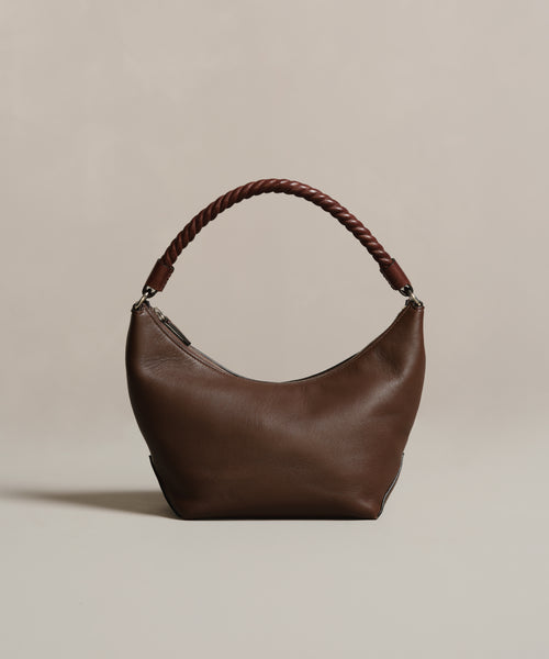 The Crescent Shoulder Bag, crafted in Italy from supple brown leather, showcases a curved silhouette and a thick twisted handle, displayed on a beige background.