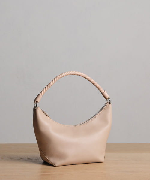 The Crescent Shoulder Bag in beige, crafted in Italy from soft leather with a braided handle, rests on a light wooden surface against a plain light gray background.