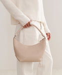 A person in a cream sweater and light pants holds the Crescent Shoulder Bag, handmade in Italy from soft beige leather, creating a refined, neutral-toned look.