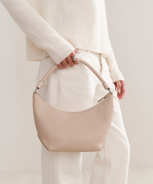 A person in a cream sweater and light pants holds the Crescent Shoulder Bag, handmade in Italy from soft beige leather, creating a refined, neutral-toned look.