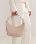 A person in a cream sweater and off-white pants holds the Crescent Shoulder Bag, handmade in Italy from soft leather, against a light neutral background.