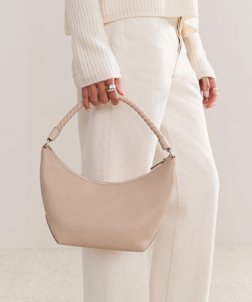 A person in a cream sweater and off-white pants holds the Crescent Shoulder Bag, handmade in Italy from soft leather, against a light neutral background.