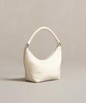 The Crescent Shoulder Bag, handmade in Italy from soft lambskin, features a thick twisted handle and minimalist design. Its cream color and elegant crescent shape stand out against a neutral beige background, highlighting its smooth texture.