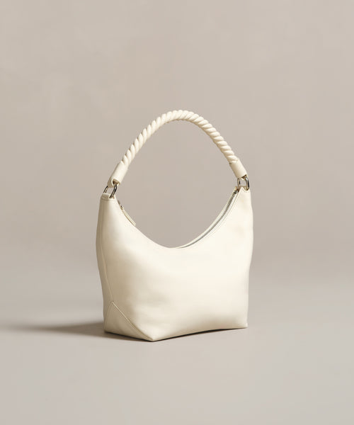 The Crescent Shoulder Bag, handmade in Italy from soft lambskin, features a thick twisted handle and minimalist design. Its cream color and elegant crescent shape stand out against a neutral beige background, highlighting its smooth texture.