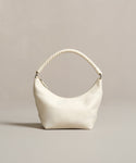 The Crescent Shoulder Bag, handmade in Italy from soft lambskin in a cream color, features a crescent shape and thick twisted handle, showcased against a plain beige backdrop.