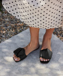 A person wearing a white skirt with black polka dots and Dani Slide black square toe sandals stands on a sidewalk near some pebbles.