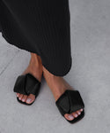 A person stands on a light gray concrete surface, wearing a long black ribbed skirt and the Dani Slide—black leather sandals with square toes and wide, folded straps. Their toenails are neatly trimmed and painted light pink.