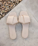 A pair of Dani Slide sandals in light beige, featuring wide folded straps and a subtle knot detail, rests on a stone surface beside small round pebbles.