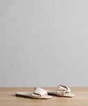 The Dani Slide, a pair of beige buttery-soft leather sandals with padded straps, sits on a light wood surface against a plain gray background.