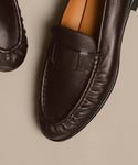 Close-up of the Leather Dylan Loafer in dark brown leather, crafted in Italy with detailed toe stitching. Displayed on a light beige surface, only the front and part of the sole are visible.