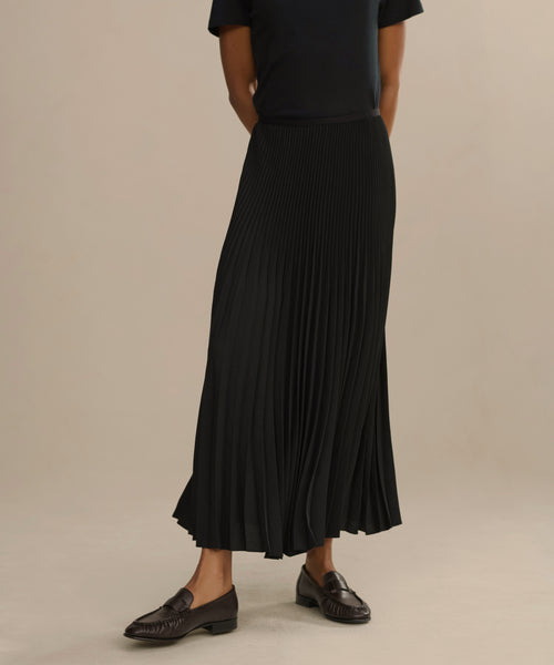 A person wearing a black short-sleeve top, black pleated maxi skirt, and the Leather Dylan Loafer stands against a plain, light background. The persons head is not visible in the image.