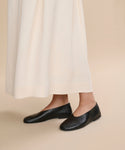 A person wearing a long cream skirt and the Leather Elodie Flat stands on a plain beige background, with only their lower legs and feet visible.