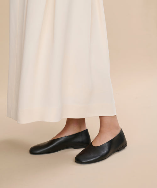 A person wearing a long cream skirt and the Leather Elodie Flat stands on a plain beige background, with only their lower legs and feet visible.