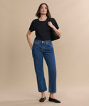 A woman stands against a beige background, wearing blue high-waisted jeans and the Leather Elodie Flat. She carries a black bag over her shoulder, her brown hair grazing her shoulders as she exudes relaxed confidence in Italian-made leather flats.