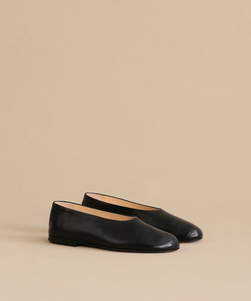 The Leather Elodie Flat, a pair of sleek black Italian-made flats with rounded toes and a feminine silhouette, is displayed on a light beige background.