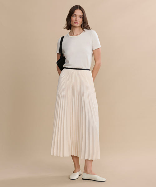 A woman poses against a beige backdrop, wearing a white short-sleeve top, cream pleated midi skirt, and the Leather Elodie Flat for an elegant touch. She carries a black shoulder bag and lets her hair fall loosely at her sides.