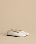 The Leather Elodie Flat, an Italian-made leather shoe with black soles, is displayed on a light beige surface against a matching background, highlighting its timeless and feminine silhouette.