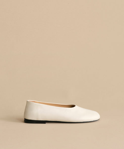 The Leather Elodie Flat is a minimalist Italian-made leather shoe with a rounded toe, black sole, and feminine silhouette, shown against a light beige background.