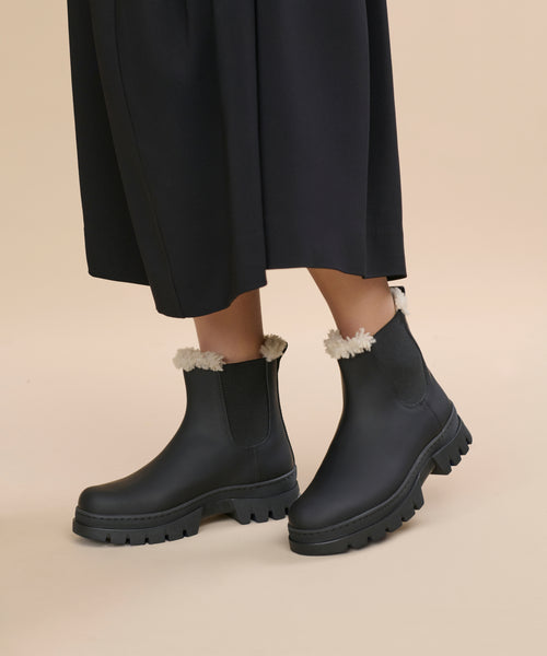 A person wearing a black skirt and Leather Highland Boot—black ankle boots with chunky soles and white shearling lining—stands on a beige background.