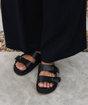 A person wearing black wide-leg pants and Hutton Slide Sandals stands on a concrete surface with small pebbles in the background—an effortlessly chic everyday staple look.