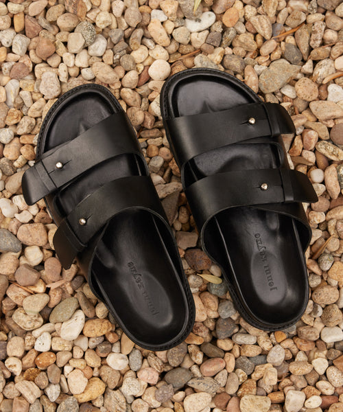 The Hutton Slide Sandal, featuring two black leather straps with metal studs, is shown side by side on light pebbles—a chic everyday staple for effortless style.