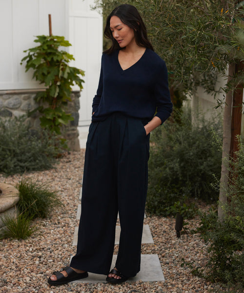 A person with long dark hair wears a navy V-neck sweater, wide-leg trousers, and the Hutton Slide Sandal—an everyday staple—while standing on a stone path in a garden surrounded by greenery and pebbles, looking down with one hand in their pocket.