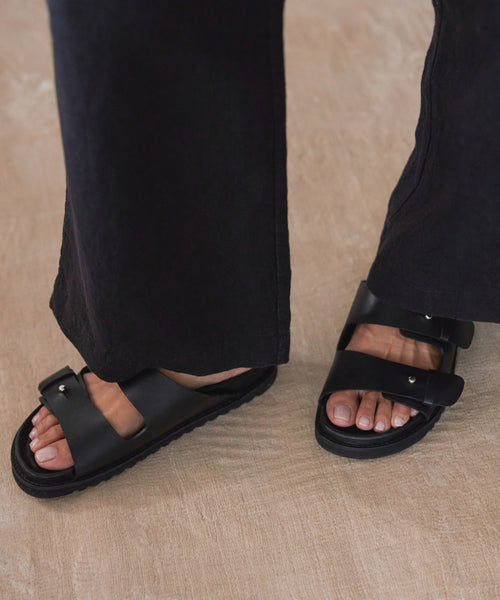 A person stands on a light floor wearing black wide-leg pants and the Hutton Slide Sandal in black leather. Only their lower legs and feet are visible, highlighting this stylish staple shoe.