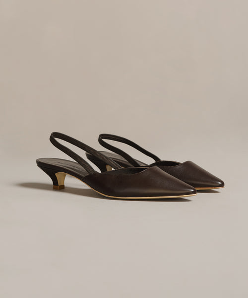 The Leather Kitten Heel features dark brown, pointed-toe leather with low heels and gold accents, crafted in Italy and shown on a neutral background.