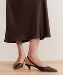 A person stands on a light wood floor wearing a dark brown midi skirt and the Leather Kitten Heel in brown, featuring pointed toes and slingback straps.