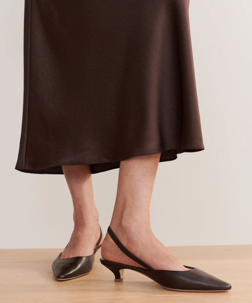 A person stands on a light wood floor wearing a dark brown midi skirt and the Leather Kitten Heel in brown, featuring pointed toes and slingback straps.
