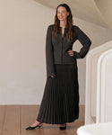 A woman with long brown hair smiles indoors, posing near a white staircase. She wears a charcoal cardigan, long pleated black skirt, and the Leather Kitten Heel, standing confidently with one hand on her hip.