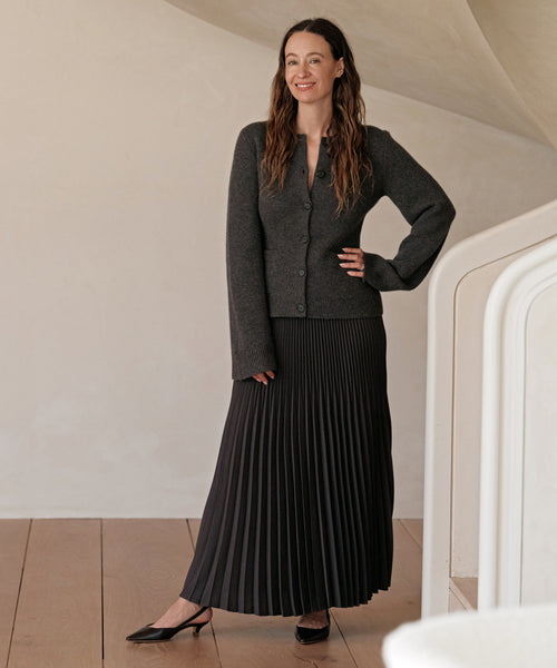 A woman with long brown hair smiles indoors, posing near a white staircase. She wears a charcoal cardigan, long pleated black skirt, and the Leather Kitten Heel, standing confidently with one hand on her hip.