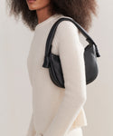 A woman with curly hair wears a cream ribbed sweater and holds the Loren Shoulder Bag, crafted in soft pebbled leather. The image, cropped from shoulders to hips against a plain background, showcases this stylish Italian-made bag.