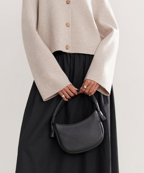A person wearing a beige long-sleeved buttoned cardigan and black skirt holds the Loren Shoulder Bag in soft pebbled leather with both hands in front of their torso; only the torso and hands are visible.